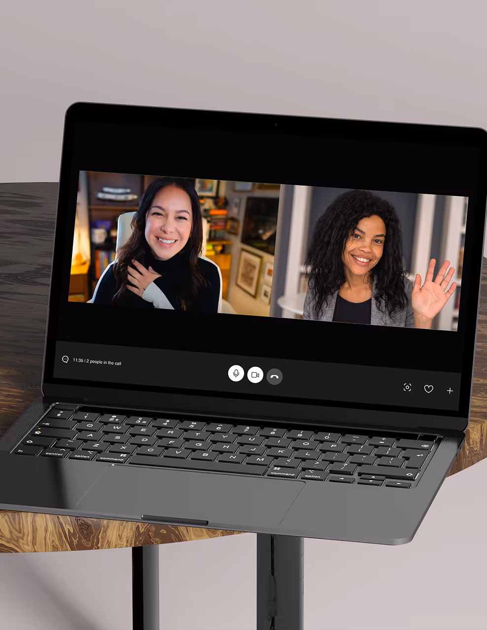 Laptop on wooden table showing video call with two smiling women, one waving and one touching chest.