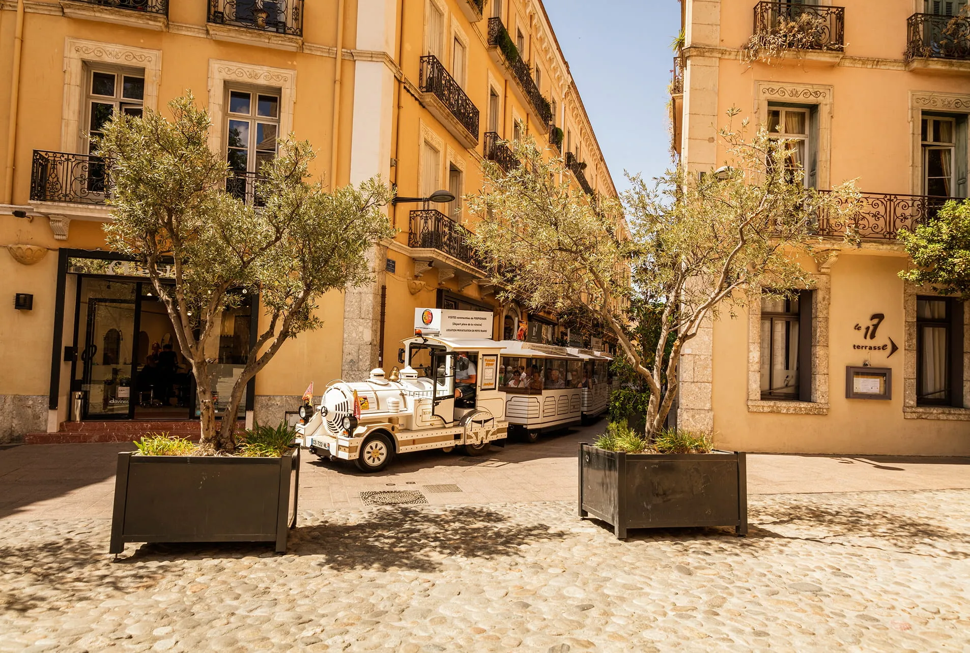 Petit train touristique blanc avec passagers circulant entre des bâtiments jaunes et des arbres en pots sur une place pavée.