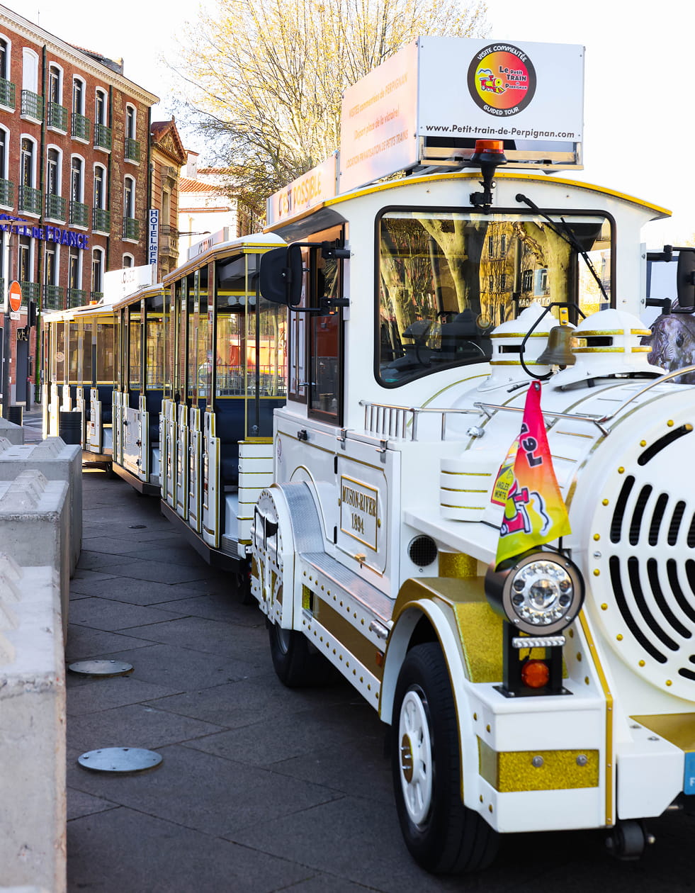 Petit train touristique blanc et or garé le long d'une rue pavée avec des bâtiments en arrière-plan à Perpignan.