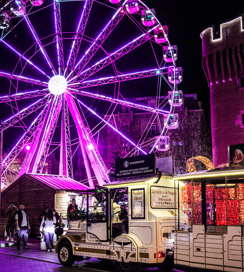 Grande roue illuminée en violet la nuit près d'un petit train touristique blanc et d'une tour ancienne éclairée.