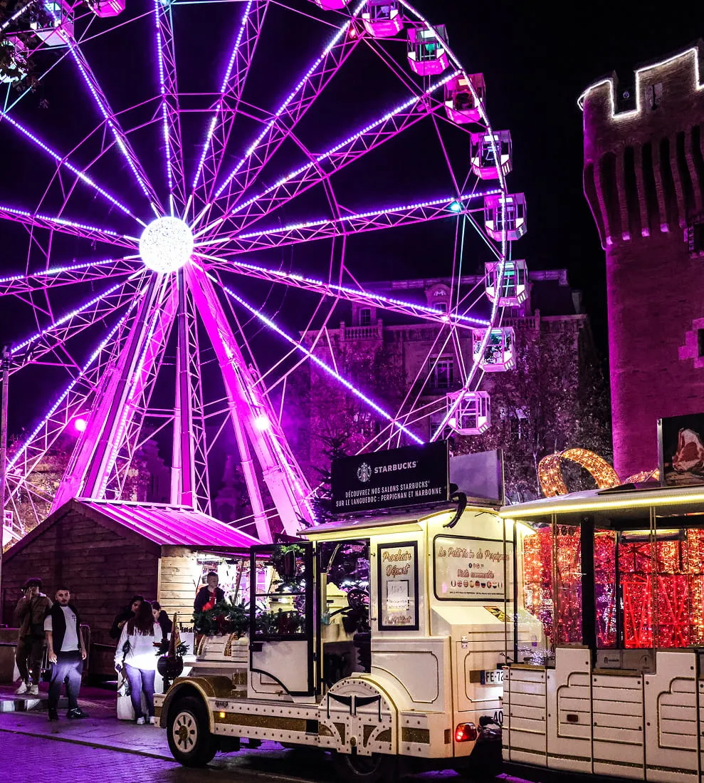 Grande roue illuminée en violet la nuit près d'un petit train touristique blanc et d'une tour ancienne éclairée.