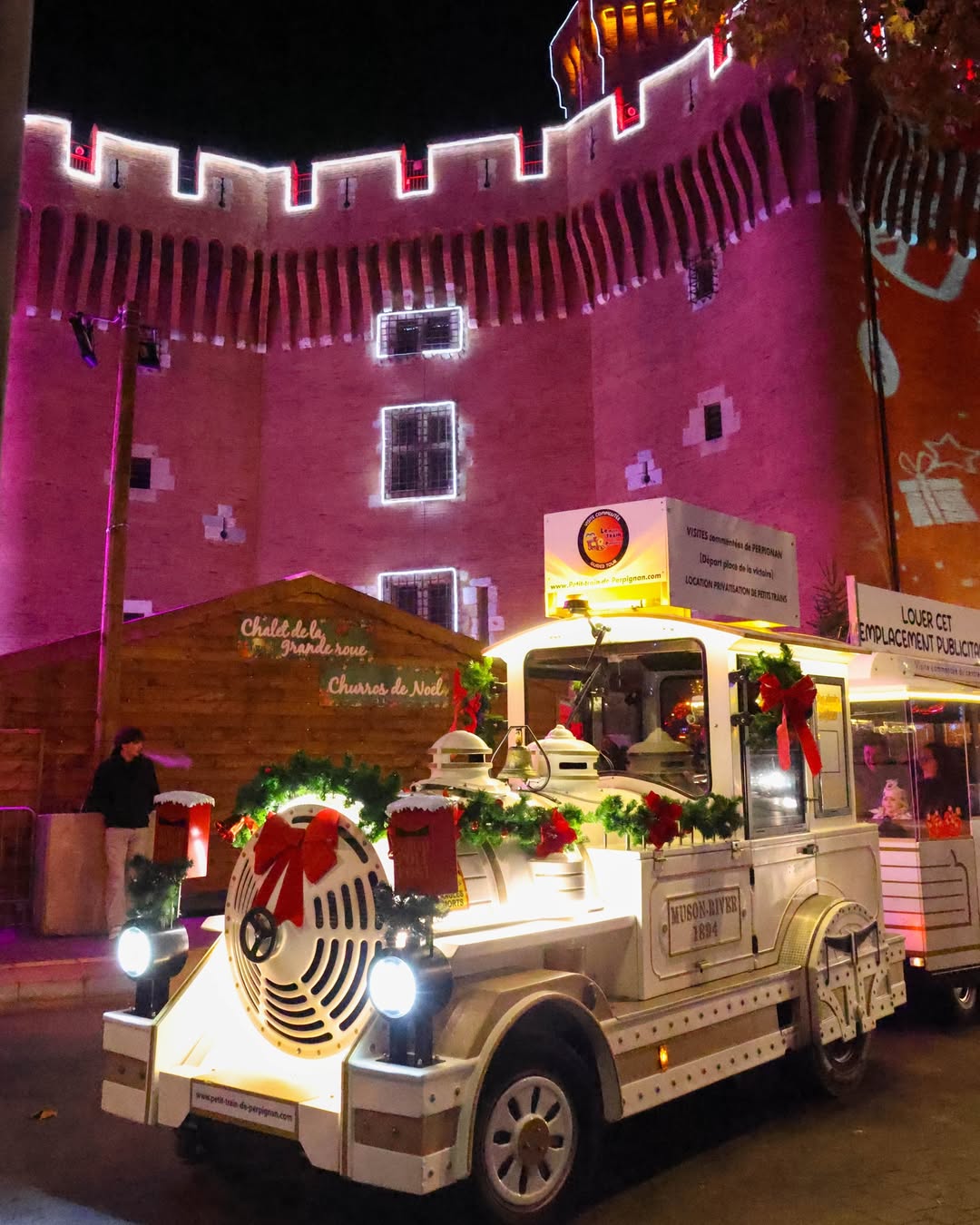 Petit train blanc décoré de guirlandes de Noël et de nœuds rouges devant un château illuminé en violet la nuit.