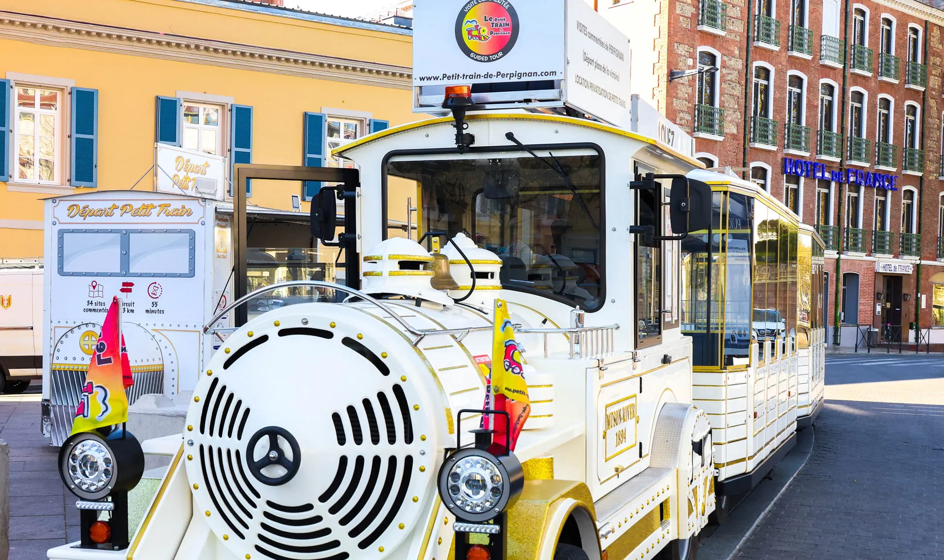 Petit train touristique blanc et or garé devant un bâtiment jaune avec fenêtres à volets bleus et un hôtel en arrière-plan.