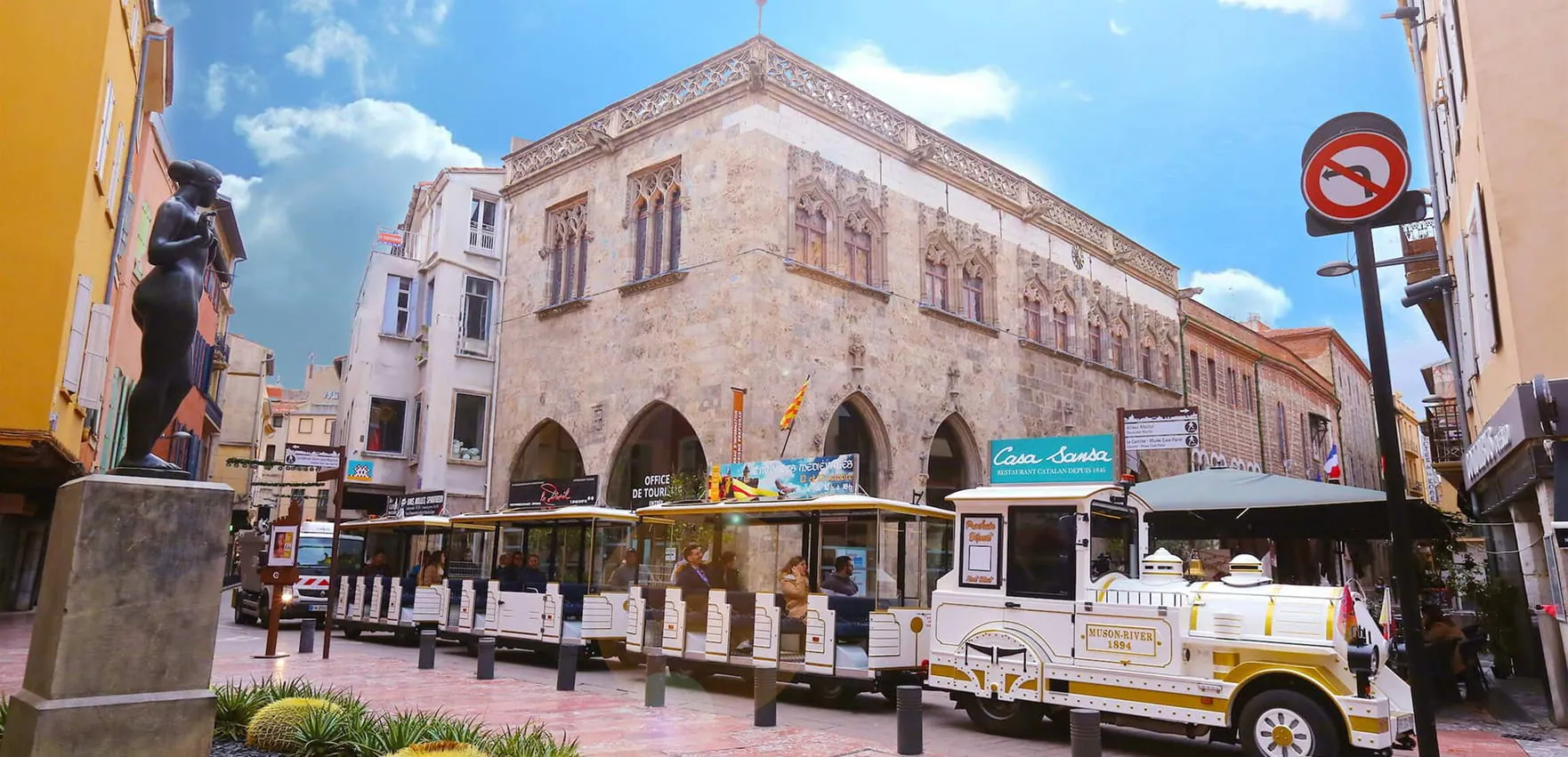 Petit train touristique blanc avec des passagers circulant devant un bâtiment historique en pierre avec des fenêtres gothiques, statue noire sur un piédestal à gauche et ciel bleu avec quelques nuages.