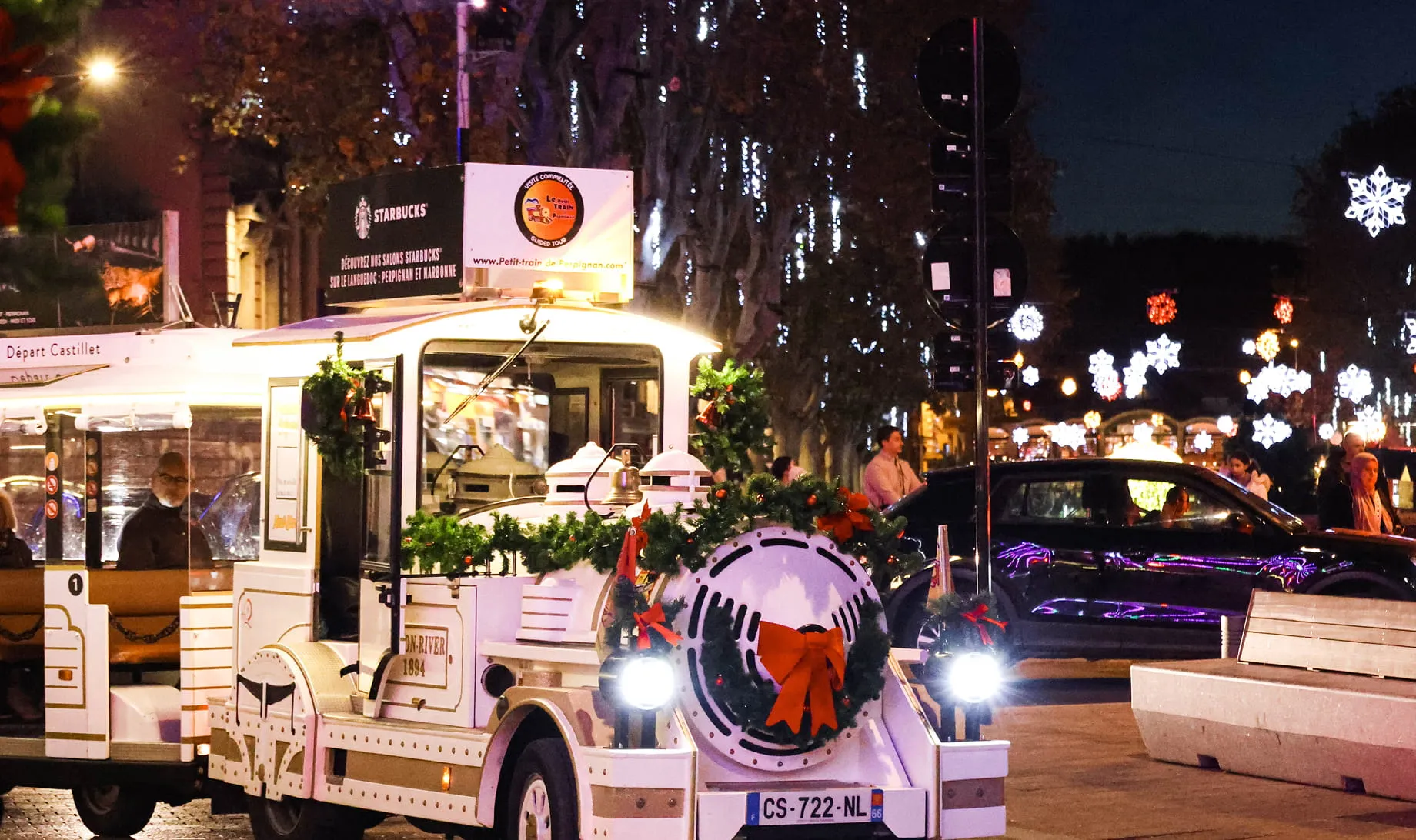 Petit train touristique blanc décoré avec des guirlandes et un grand nœud rouge, roulant de nuit avec des décorations lumineuses en forme de flocons autour.