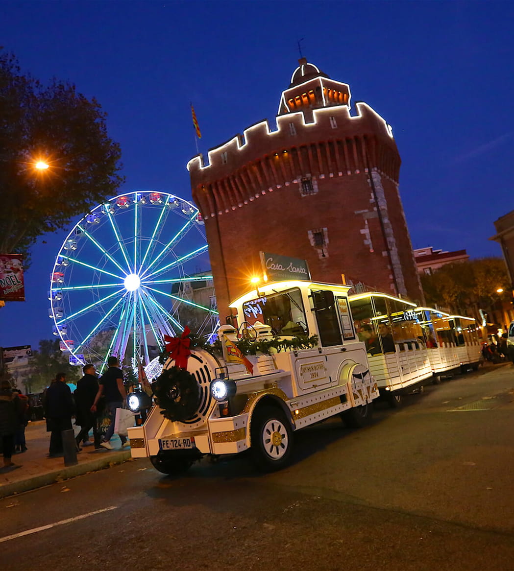 Train touristique blanc illuminé décoré pour Noël passant devant une grande roue lumineuse et une tour historique au crépuscule.