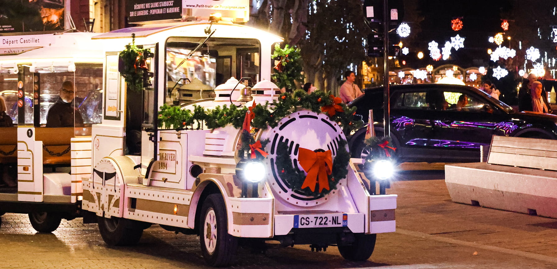 Petit train touristique blanc décoré avec des guirlandes vertes et un grand nœud rouge, circulant en ville la nuit avec des lumières festives en arrière-plan.