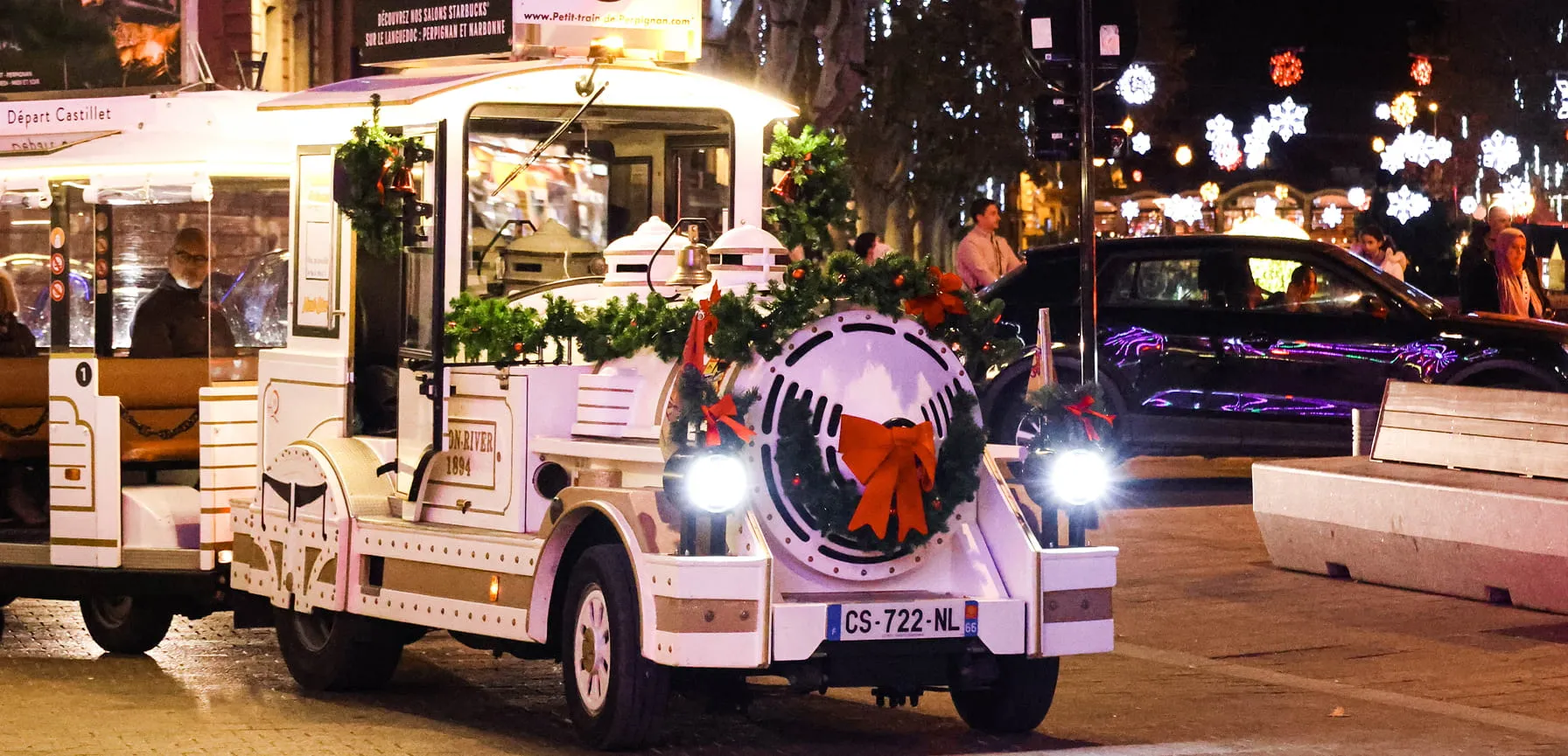 Petit train touristique blanc décoré avec des guirlandes vertes et un grand nœud rouge, circulant en ville la nuit avec des lumières festives en arrière-plan.