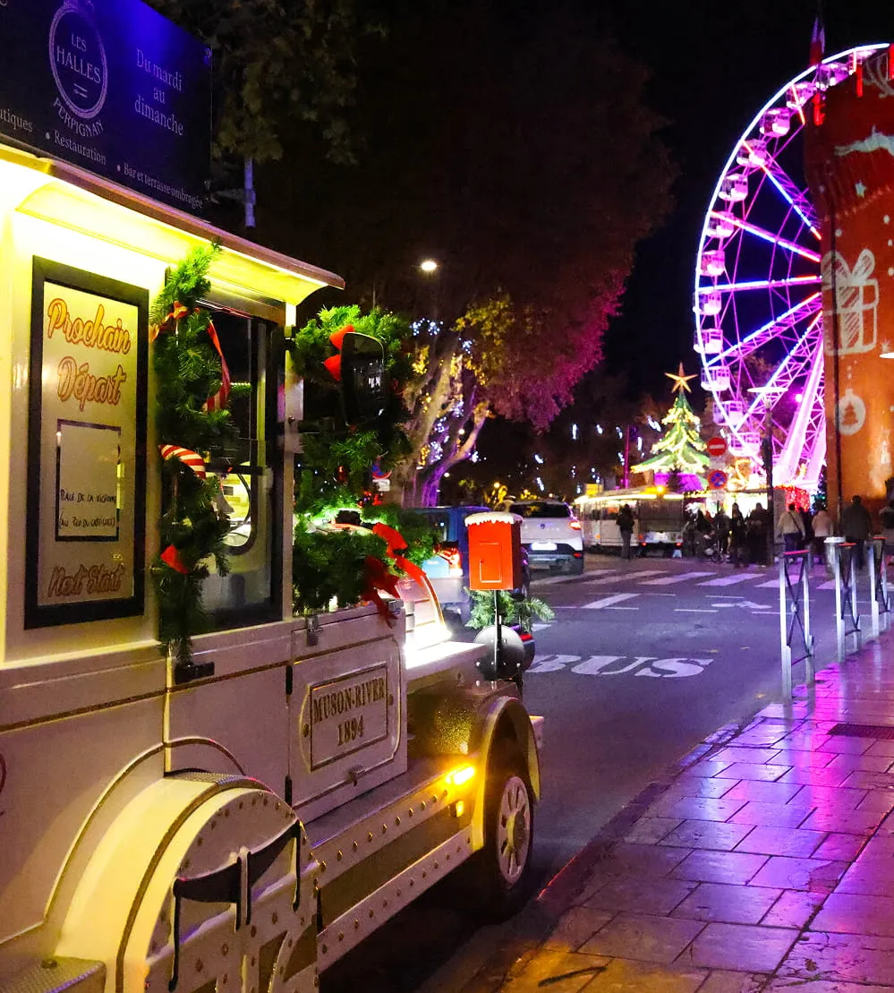 Train touristique décoré de guirlandes de Noël garé près d'une grande roue illuminée et d'un sapin de Noël dans une rue animée la nuit.