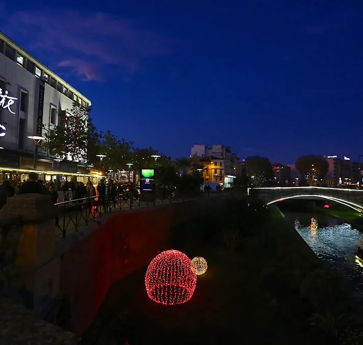 Scène nocturne d'une ville avec des sphères lumineuses rouges et blanches installées près d'une rivière sous un pont éclairé.