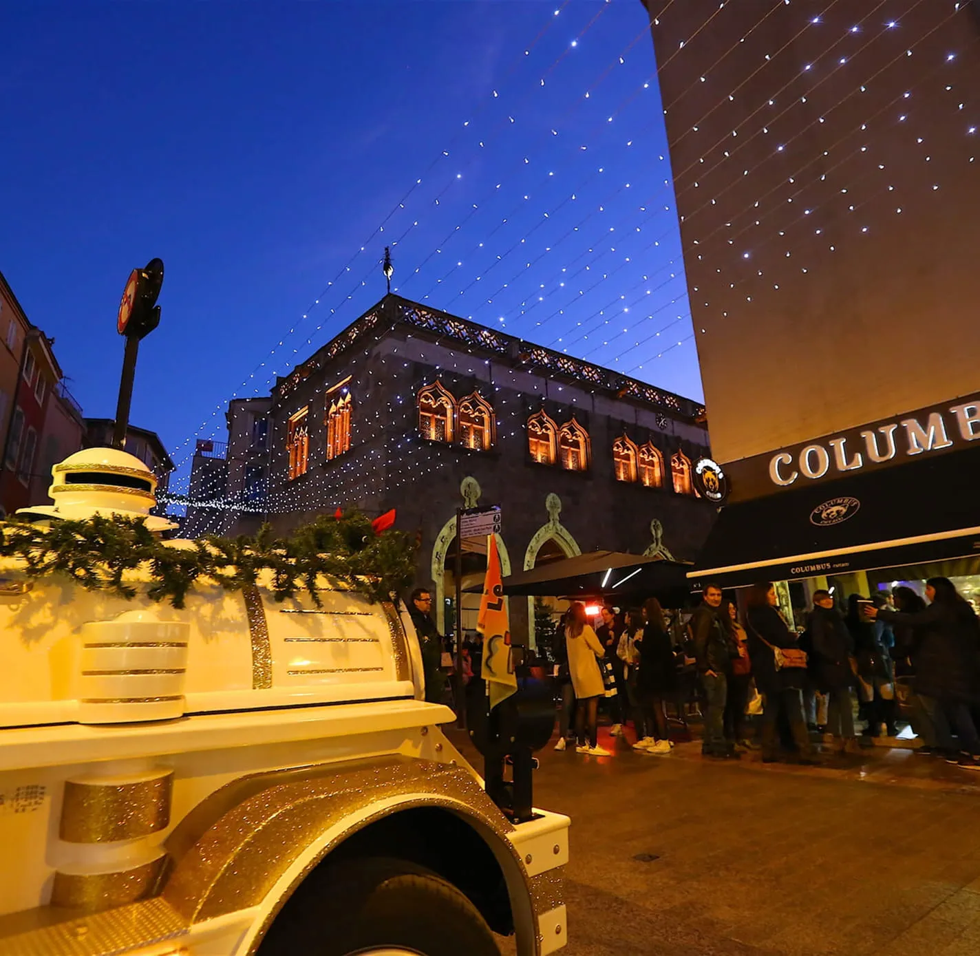 Scène nocturne d'une rue décorée de guirlandes lumineuses avec un petit train blanc et des personnes rassemblées devant un magasin Columbus Pizza.