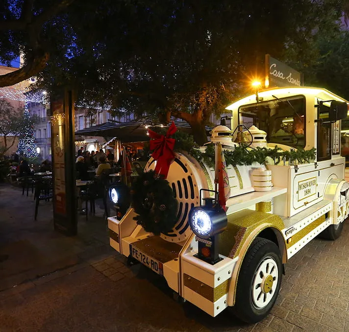 Petit train touristique décoré avec des guirlandes et une couronne de Noël dans une rue animée le soir.