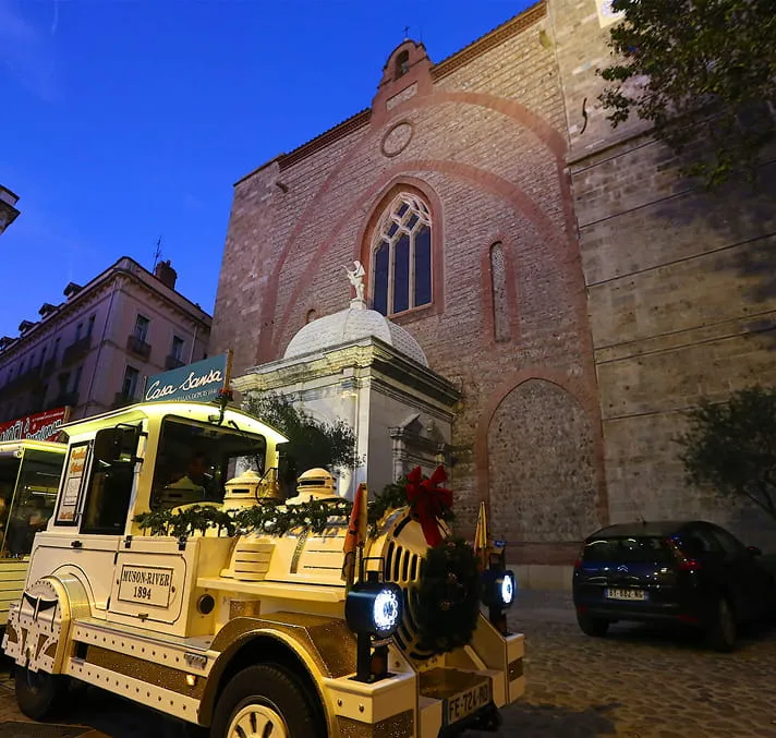 Petit train touristique décoré pour Noël garé devant un ancien bâtiment en pierre au crépuscule.