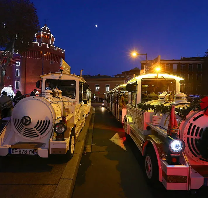 Deux petits trains touristiques blancs illuminés stationnés de chaque côté d'une rue la nuit avec un ciel bleu foncé et un bâtiment historique en arrière-plan.