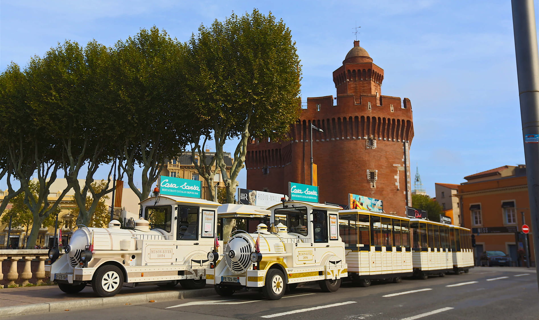 Petit train touristique blanc garé près d'un bâtiment en brique rouge en forme de tour, avec des arbres feuillus à proximité.