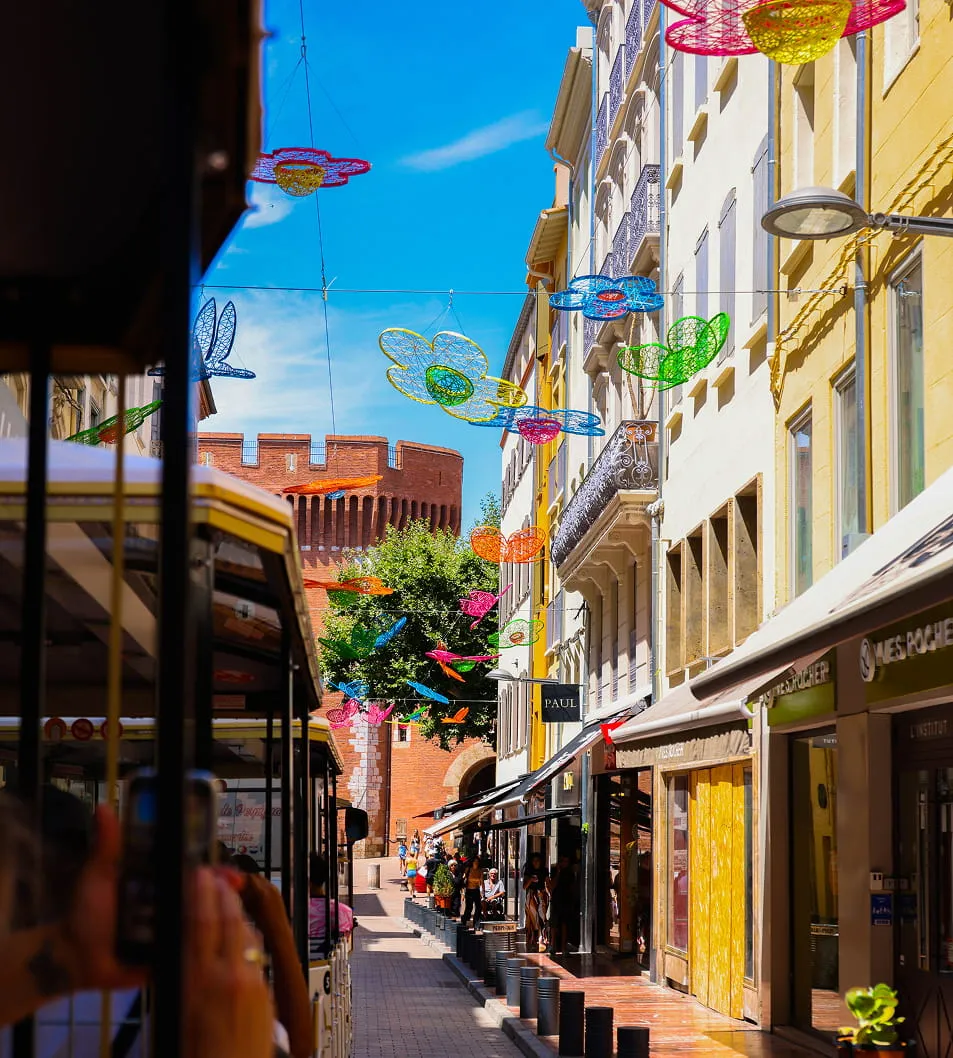 Quartier piétonnier avec des décorations florales colorées suspendues, des bâtiments aux vitrines et une tour en brique au fond sous un ciel bleu.