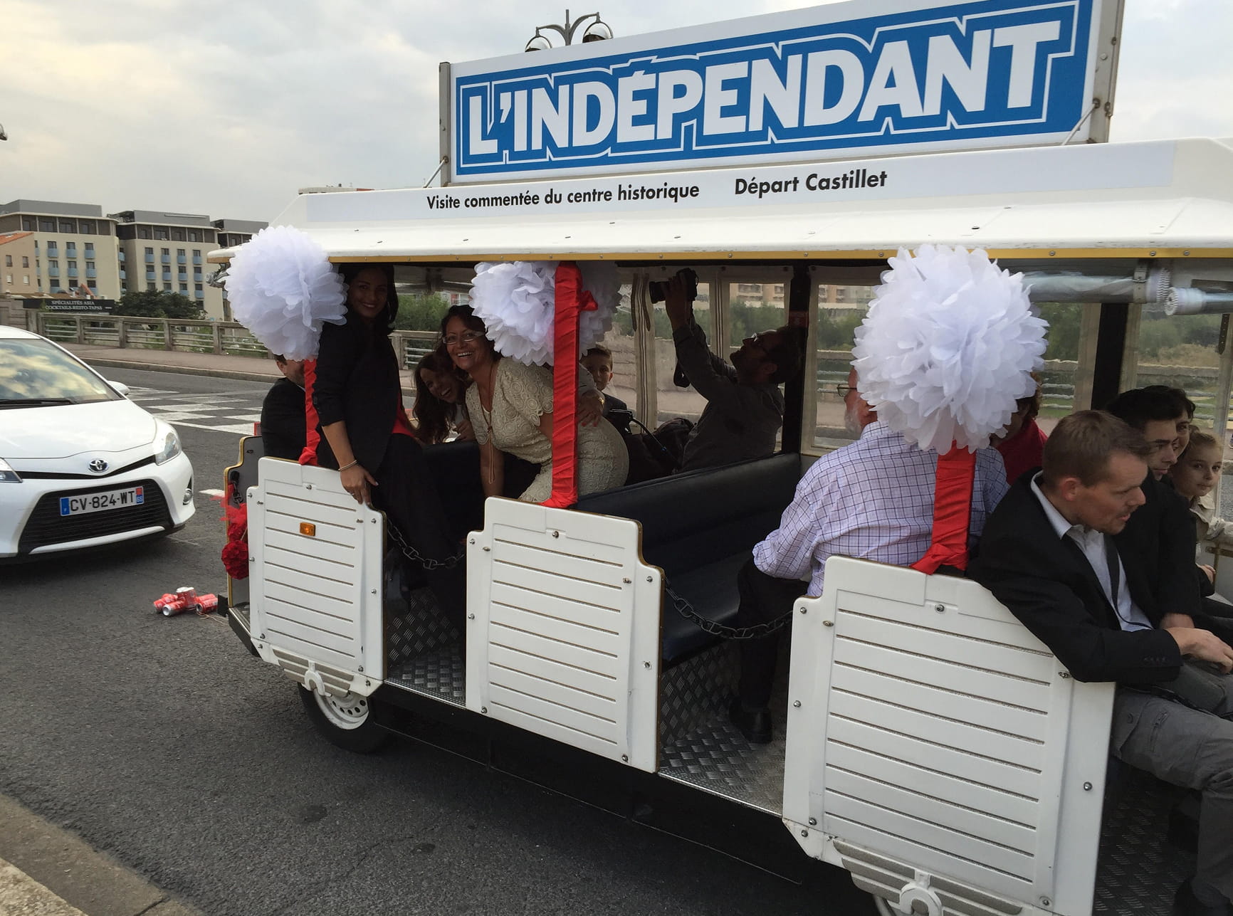 Un petit groupe de personnes assises dans un véhicule touristique ouvert décoré de pompons blancs, avec un panneau L'INDÉPENDANT sur le toit.