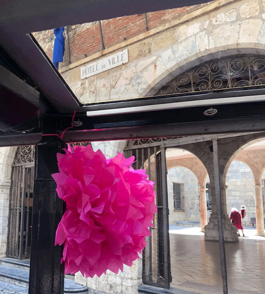 Pompon rose suspendu à une structure noire avec en arrière-plan une façade en pierre avec une enseigne Hôtel de Ville et des arches où deux personnes marchent.