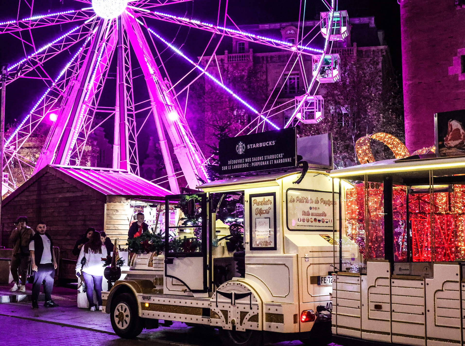 Train touristique blanc décoré avec des guirlandes lumineuses près d'une grande roue illuminée en violet la nuit.