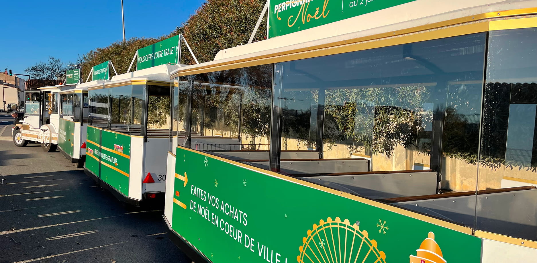Petit train touristique blanc et vert garé sur une route avec des panneaux publicitaires verts pour les achats de Noël en centre-ville.
