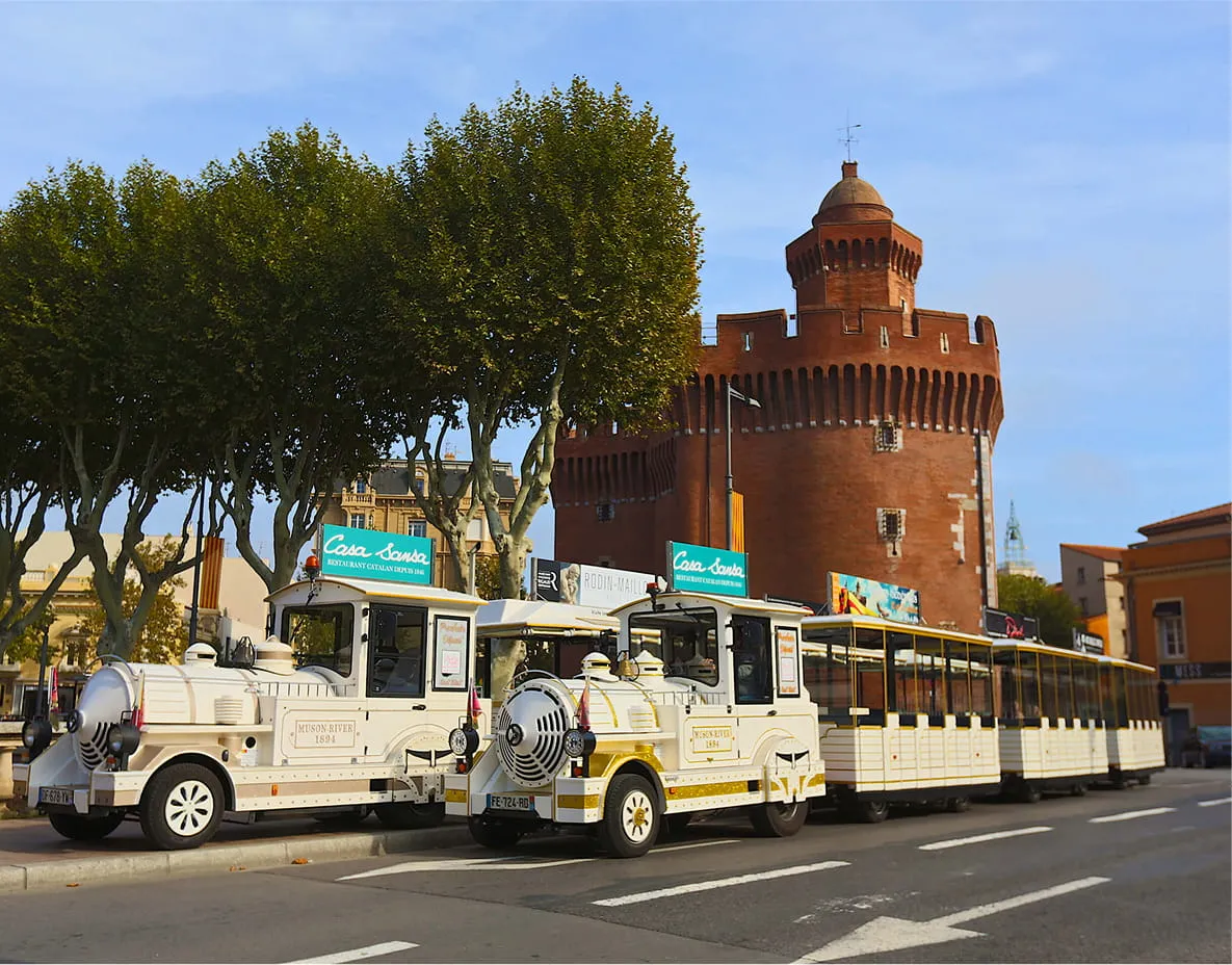 Petit train touristique blanc stationné devant une tour en brique rouge avec des arbres verts autour.