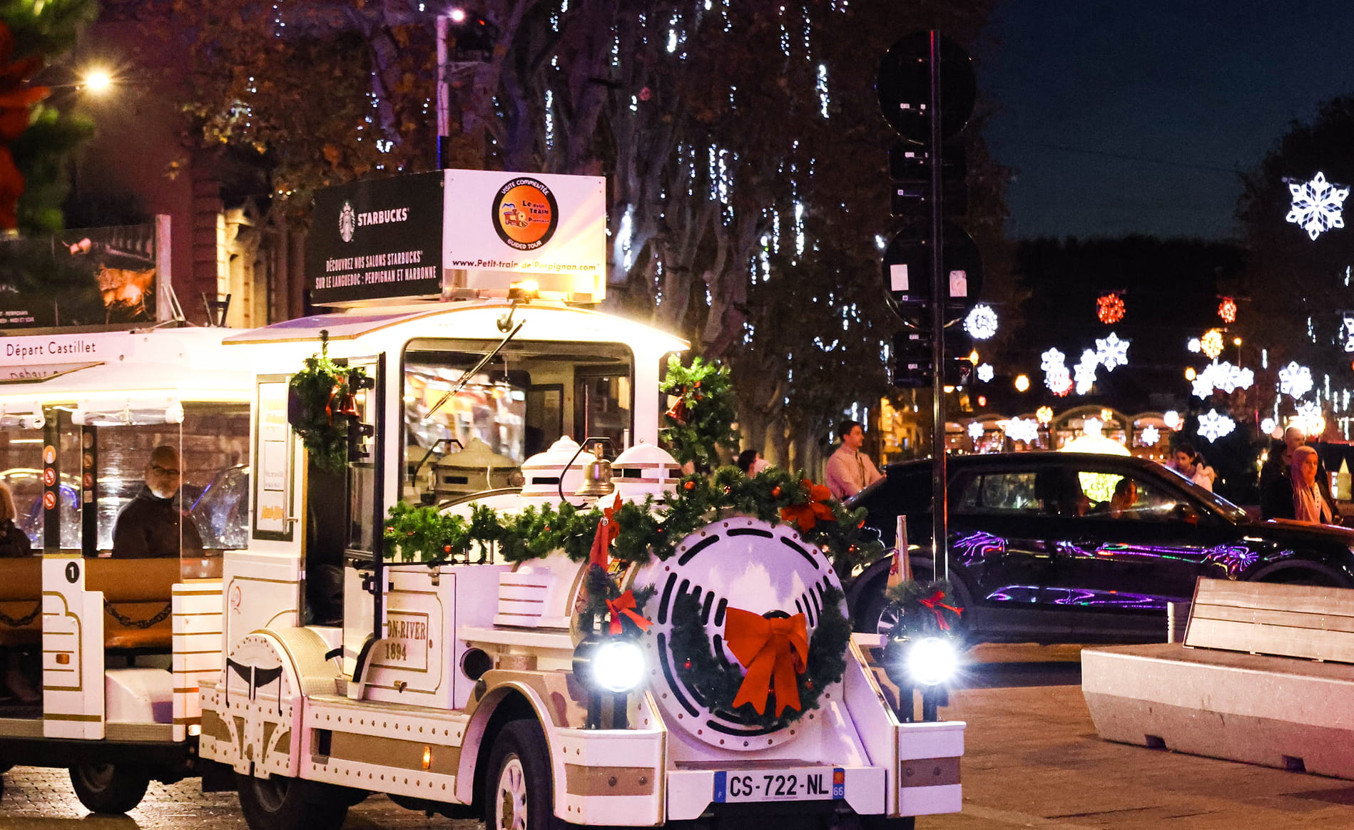 Petit train touristique blanc décoré de guirlandes et d'un grand nœud rouge, circulant de nuit sous des lumières de fête en forme de flocons.