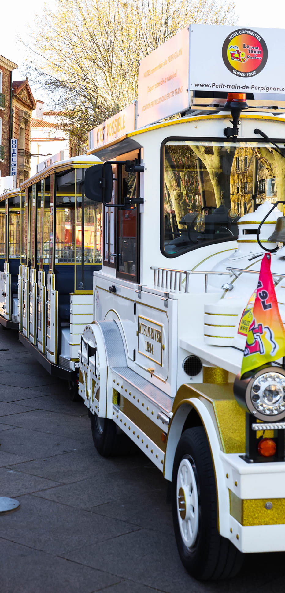 Petit train touristique blanc et doré garé en ville avec arbres en arrière-plan.