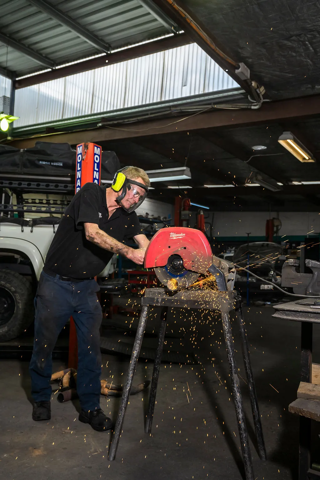 Man wearing ear protection and safety glasses using a red Milwaukee cut-off saw with sparks flying in a workshop.
