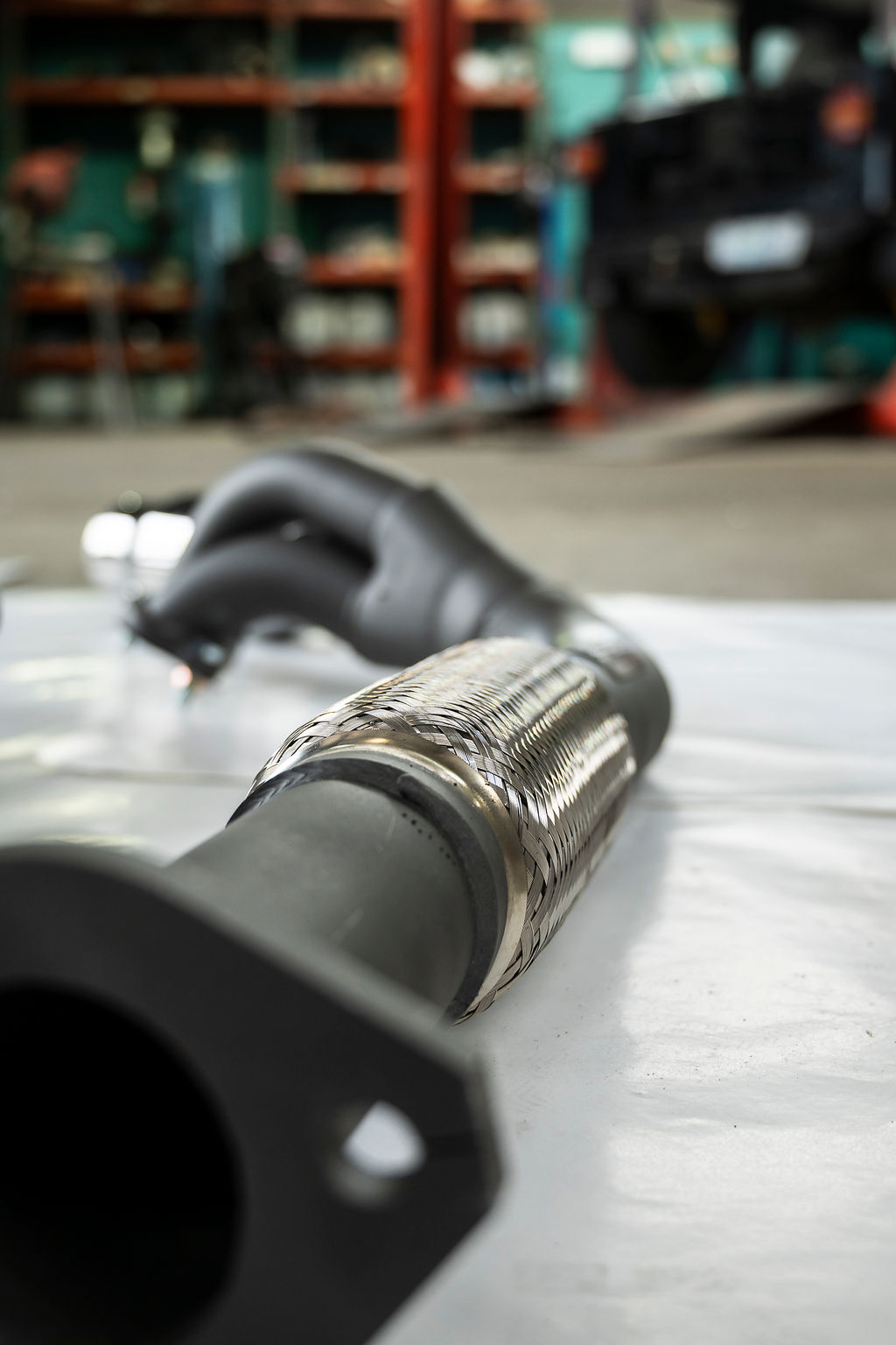 Close-up of a metallic flexible exhaust pipe on a white surface with a blurred workshop background.