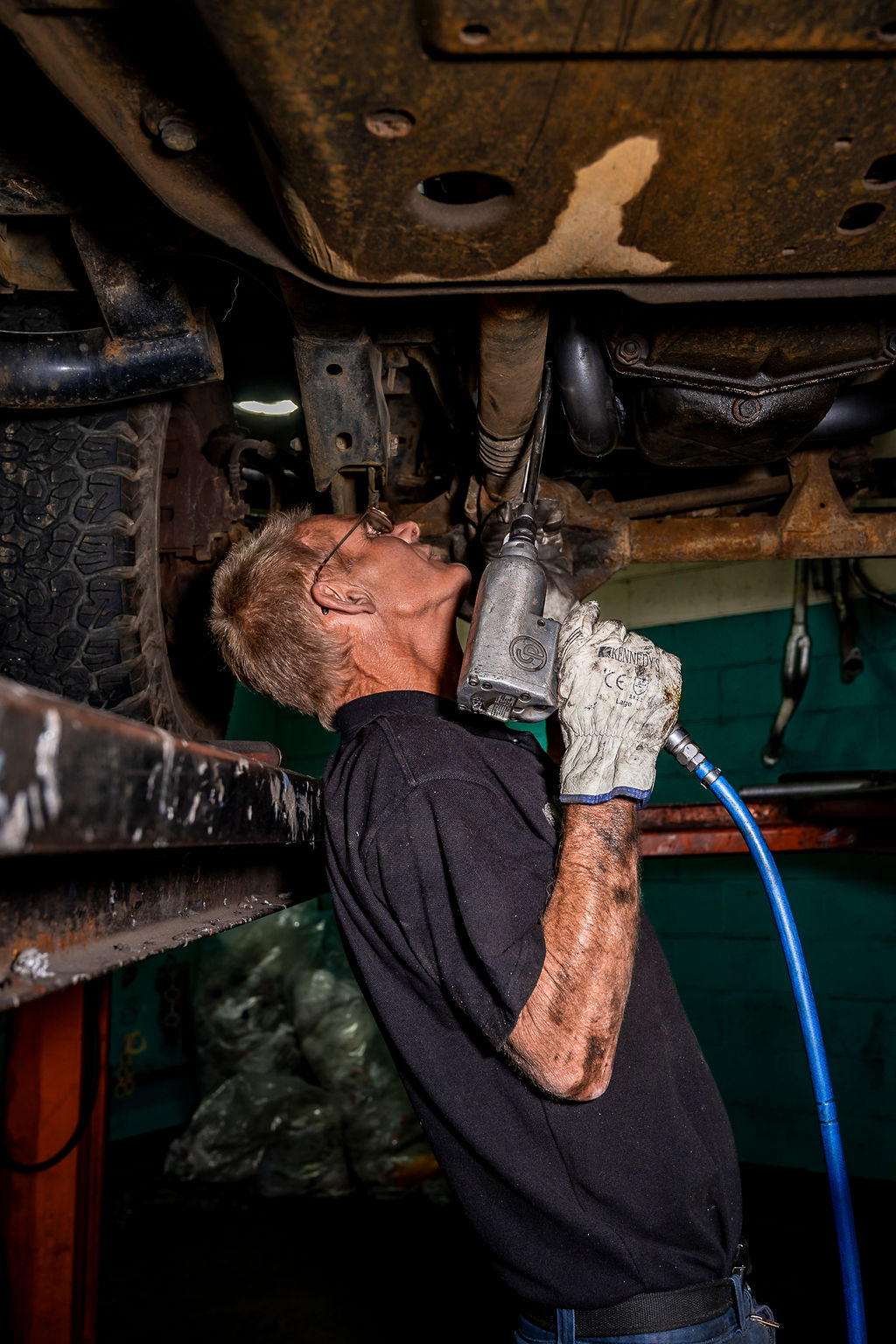 Mechanic wearing gloves and glasses using a pneumatic tool to work under a vehicle on a lift.