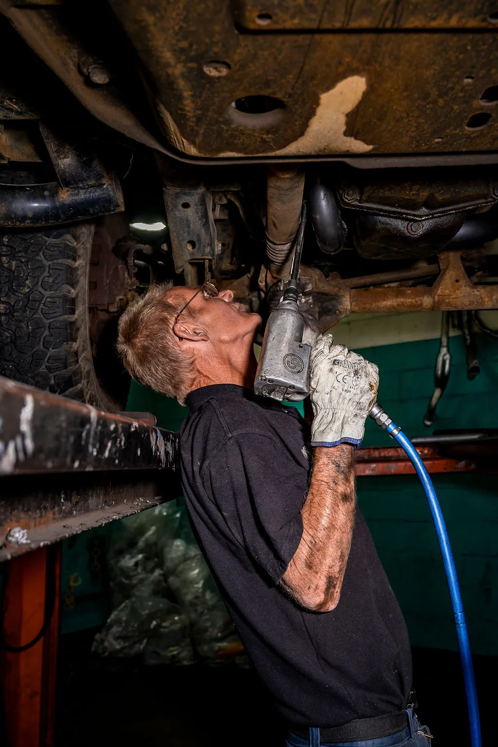 Mechanic wearing gloves and glasses using a pneumatic tool to work under a vehicle on a lift.