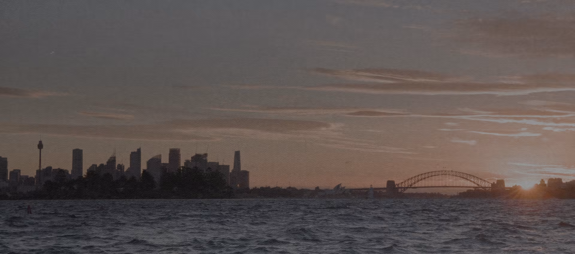 Sydney skyline at sunset with the Harbour Bridge and the Opera House silhouette over water.