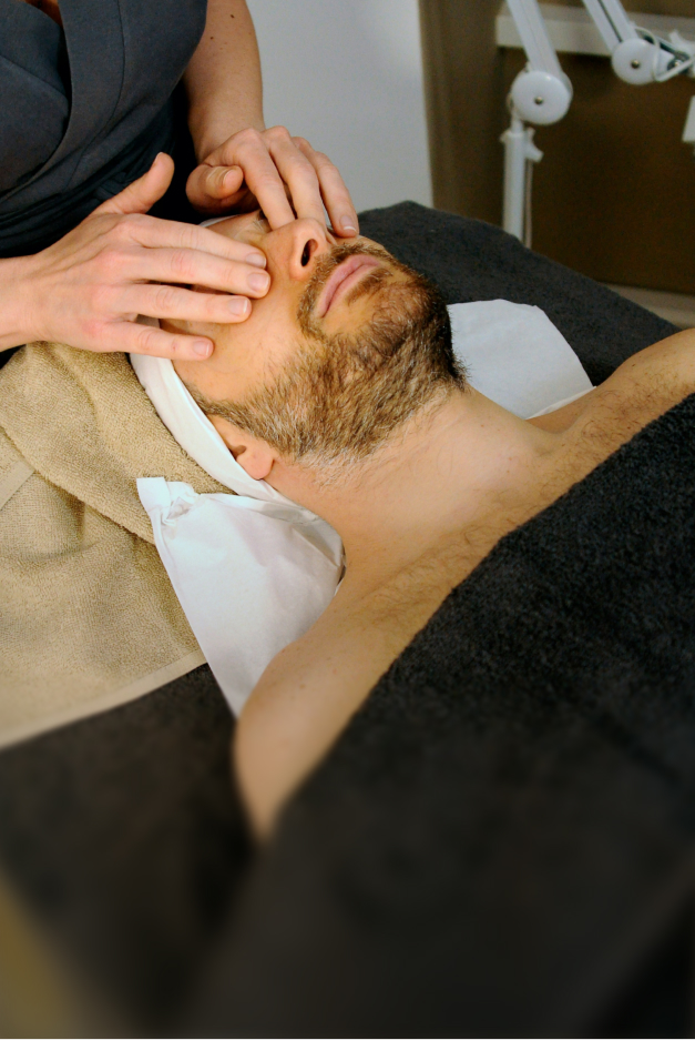 Man with beard receiving a facial massage while lying on a spa bed with a black towel.