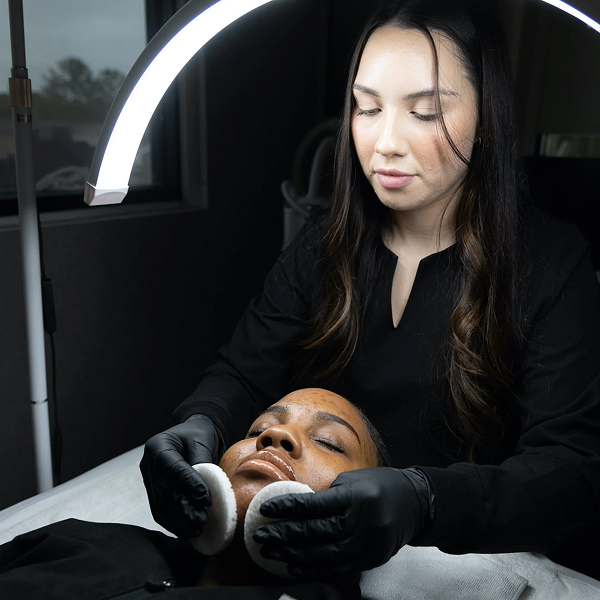 Esthetician wearing black gloves cleans a client's face under a ring light in a skincare treatment.