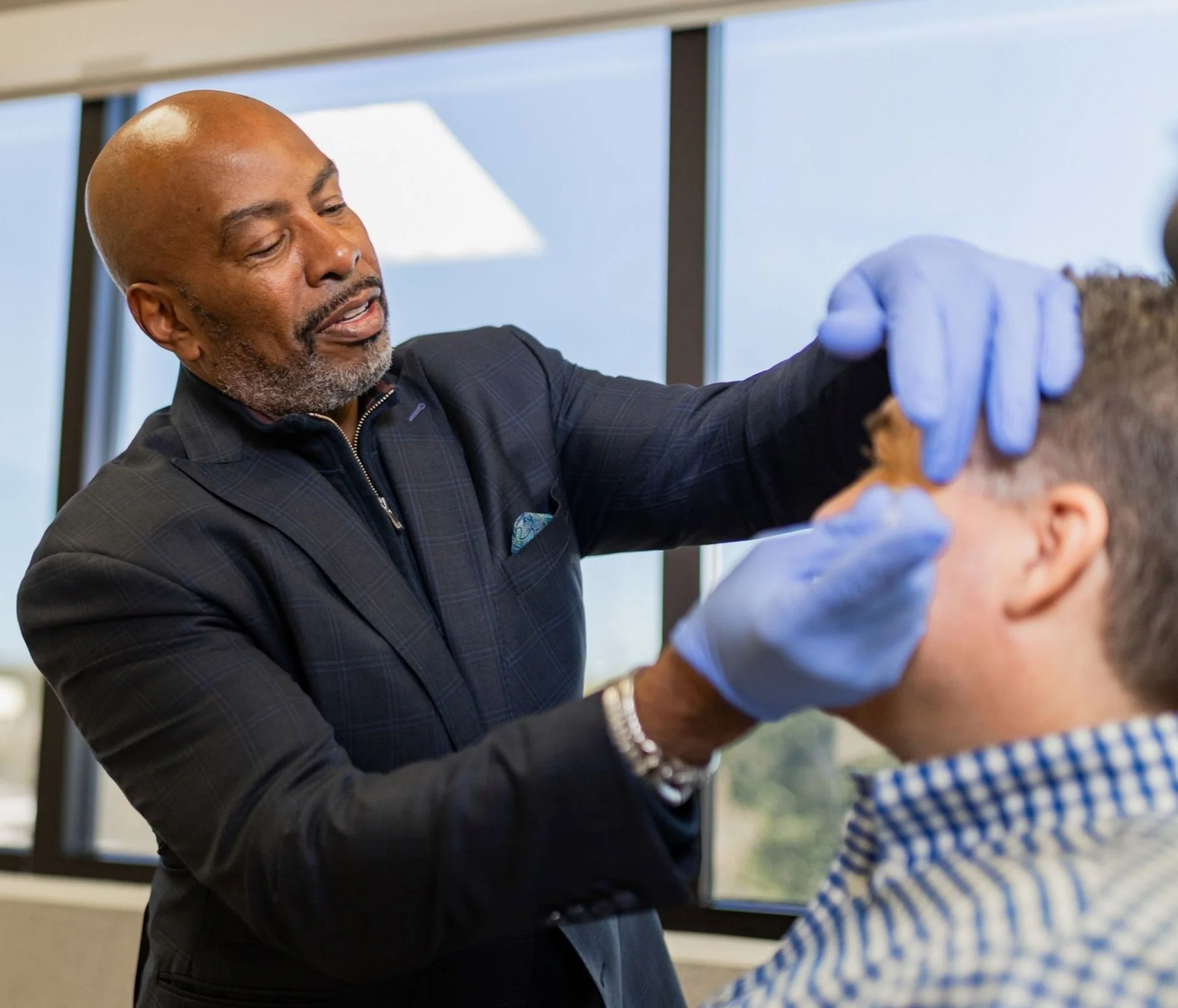 Man wearing blue gloves examining another man's face near a window in a clinical setting.