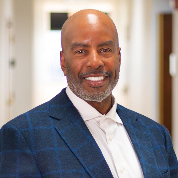 Smiling bald man with beard wearing a blue checkered blazer and white shirt in a bright hallway.