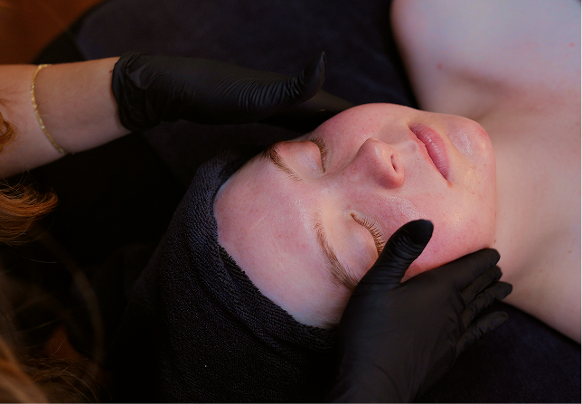 Woman with towel on head receiving a facial massage from hands wearing black gloves.