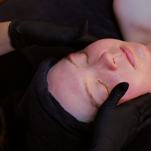 Person with eyes closed receiving facial treatment from hands wearing black gloves and a black headband.