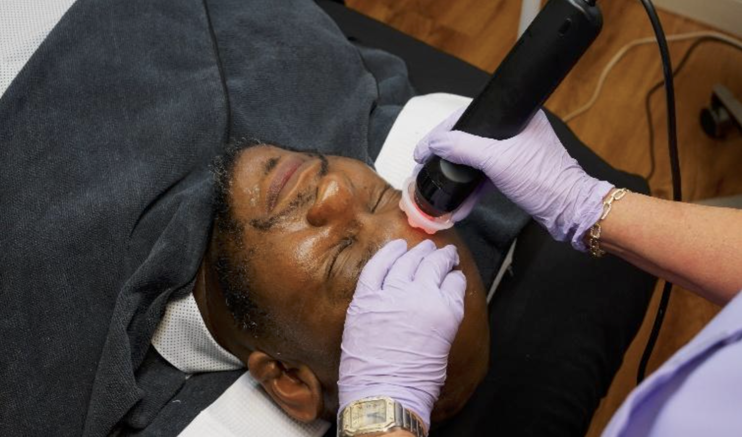 Esthetician wearing black gloves cleans a client's face under a ring light in a skincare treatment.