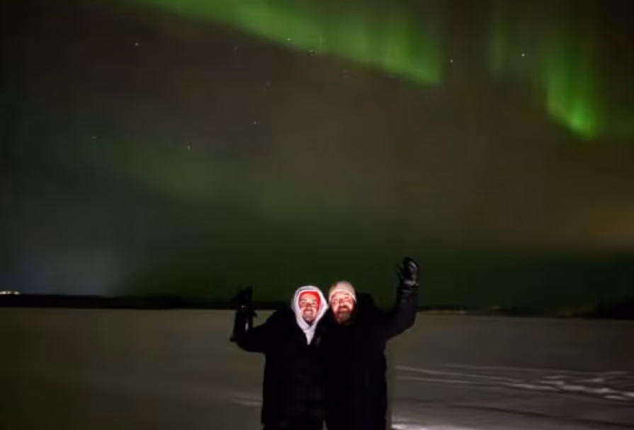 Two people in winter clothing standing on snow at night waving under bright green northern lights in the sky.