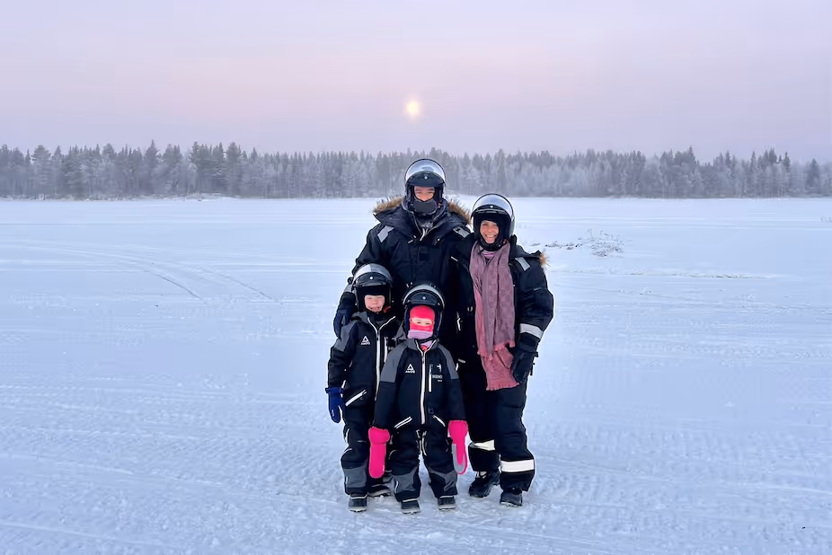 Family of four dressed in black winter gear and helmets standing on a snowy landscape with a forest and pale sky in the background.