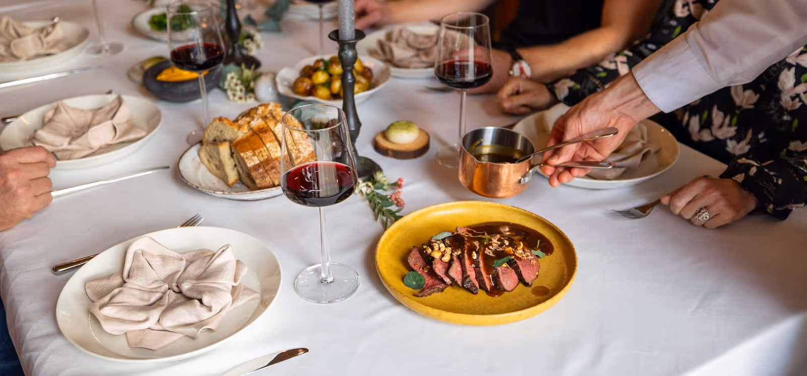 Plate of sliced steak with sauce being served on a white tablecloth with wine glasses, bread, and other dishes around.