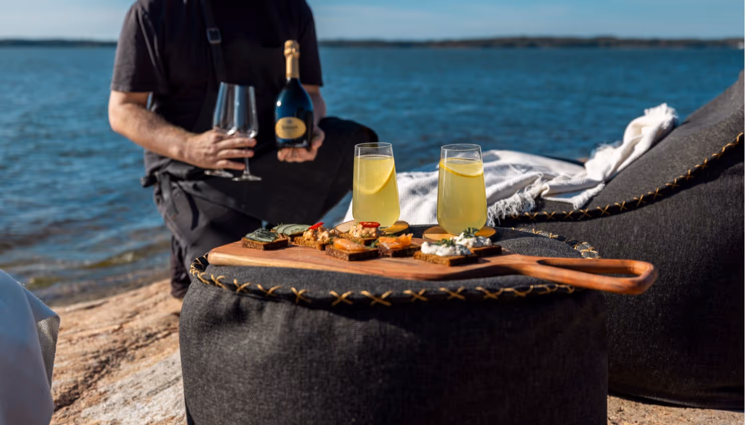 Person holding a bottle of sparkling wine and two empty wine glasses near a lake, with a wooden board of assorted appetizers and two lemon drinks on a black cushion.