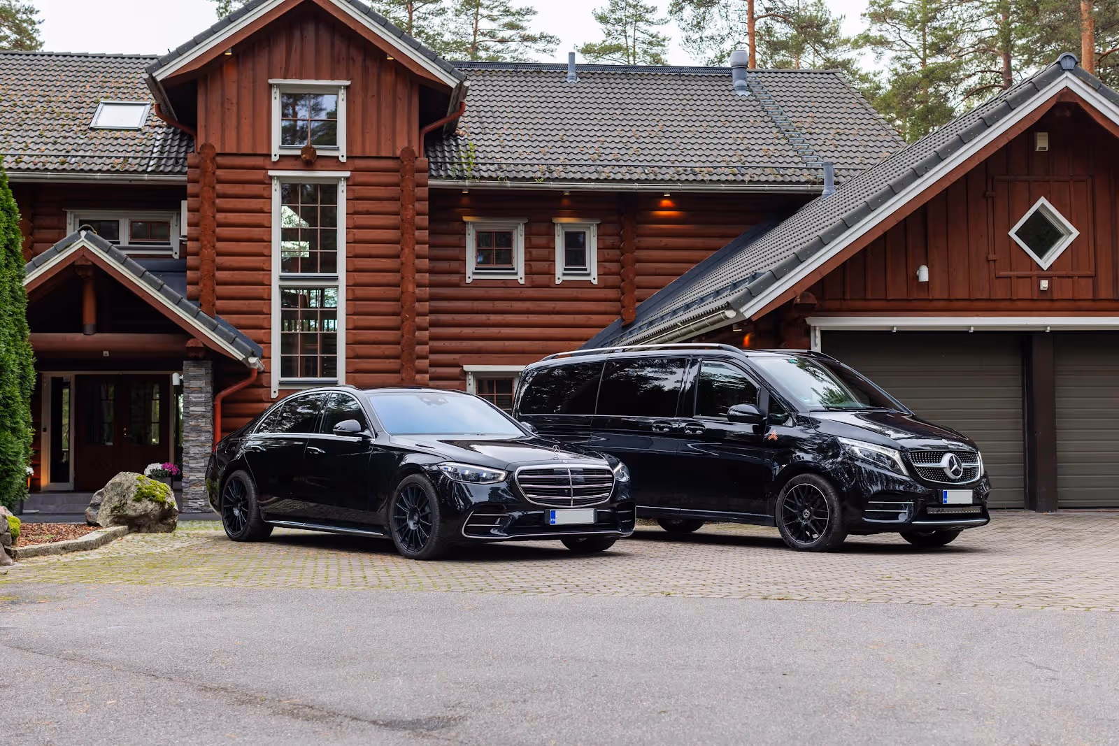 Black Mercedes-Benz sedan and black Mercedes-Benz van parked on driveway in front of a large wooden house with a garage.