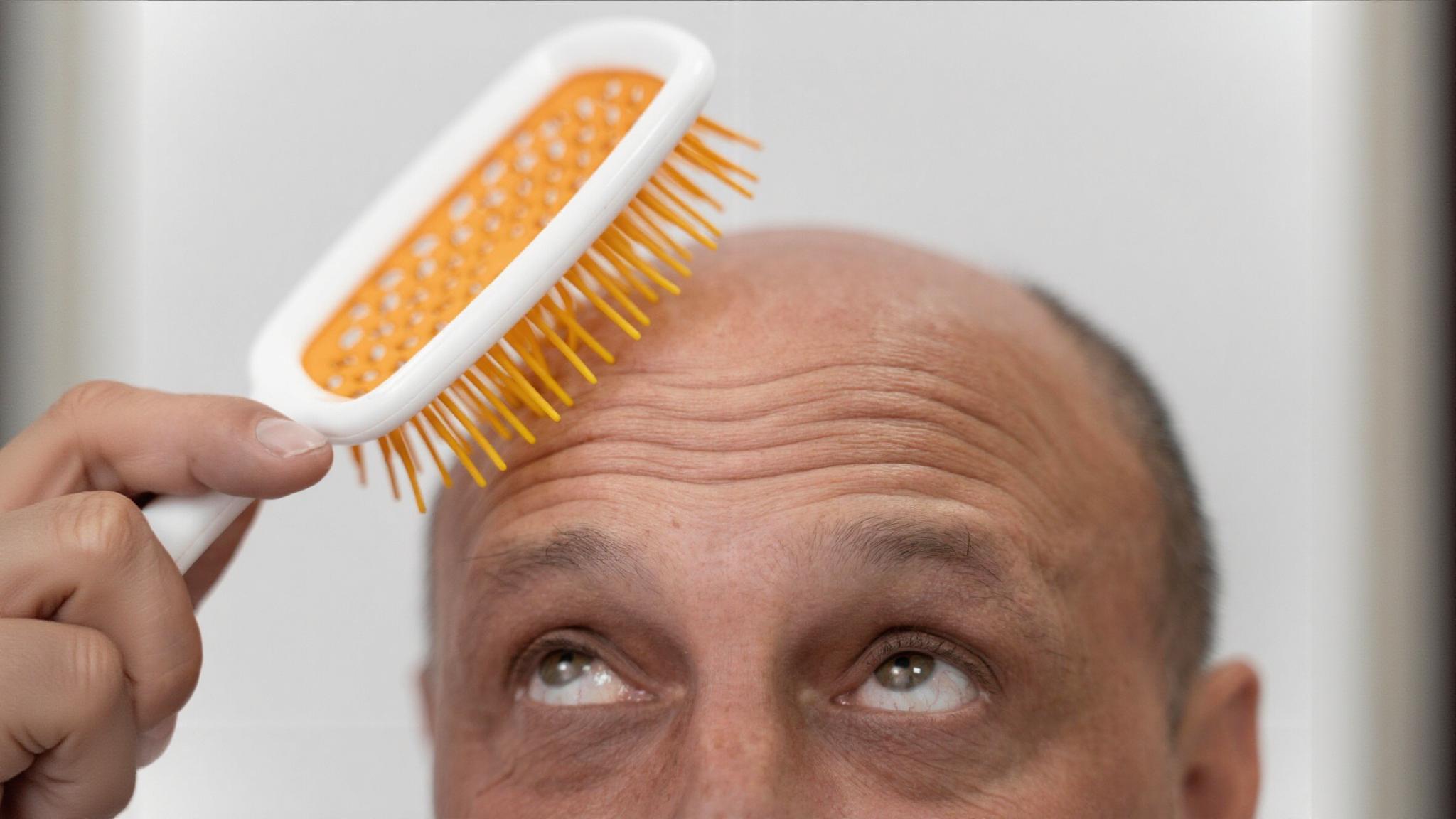 Close-up of a balding man looking up while holding an orange and white hairbrush above his head.