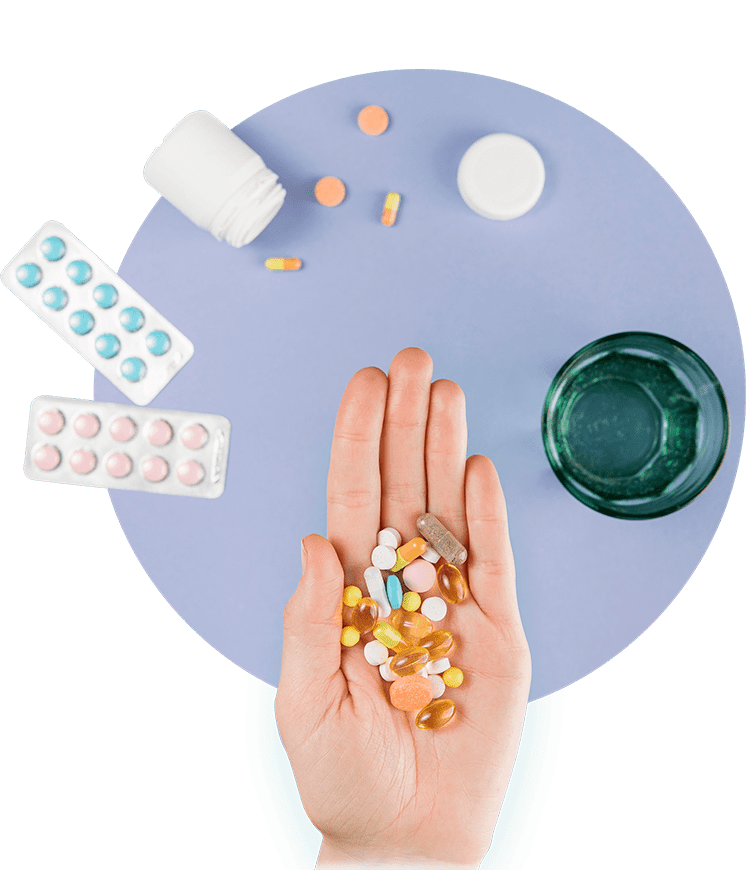 Hand holding a variety of colorful pills and capsules above a purple surface with pill bottles, blister packs, and a glass of water.