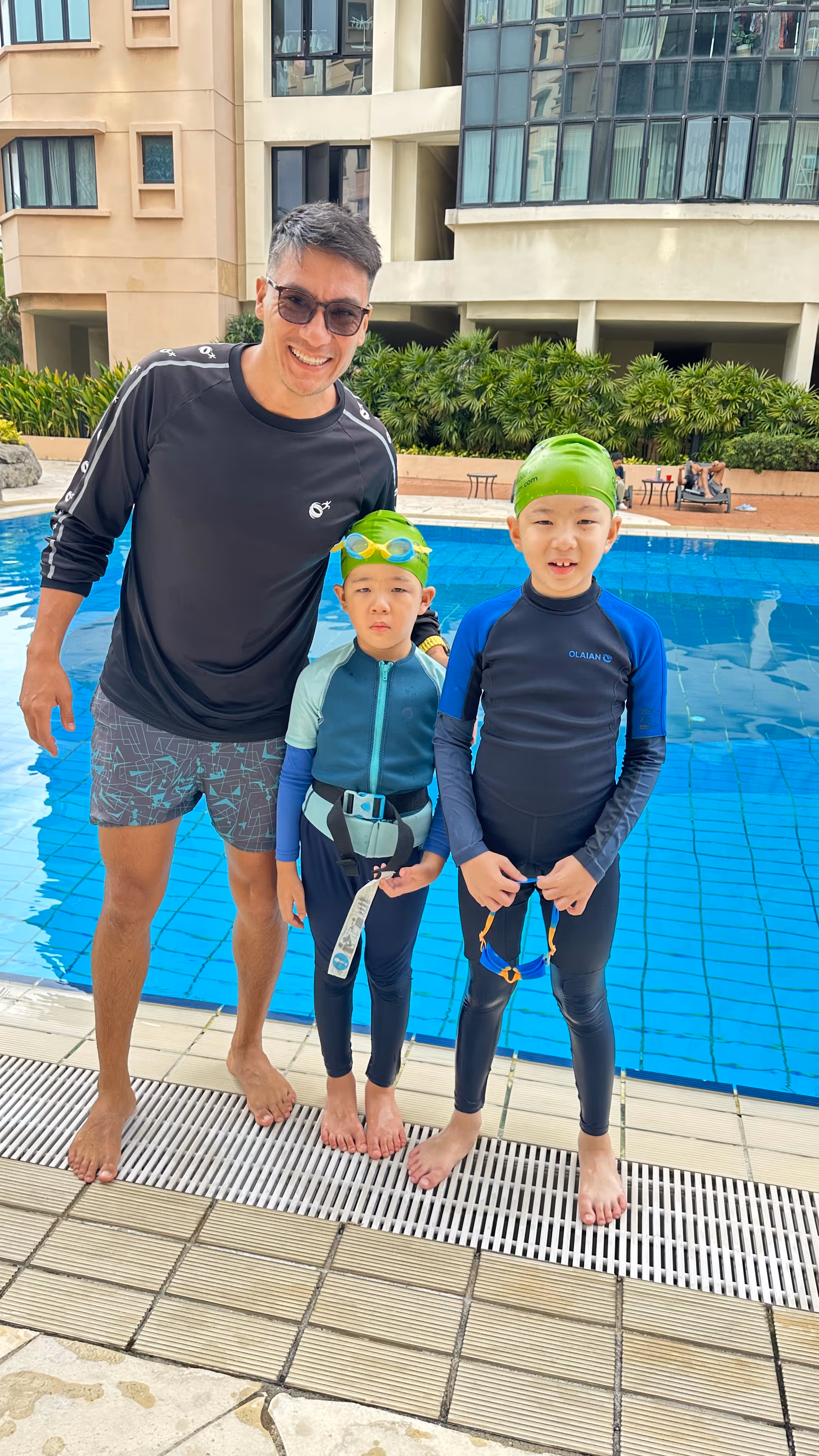 Man standing in a pool holding a smiling child wearing blue swimwear and goggles, both raising one arm in celebration.