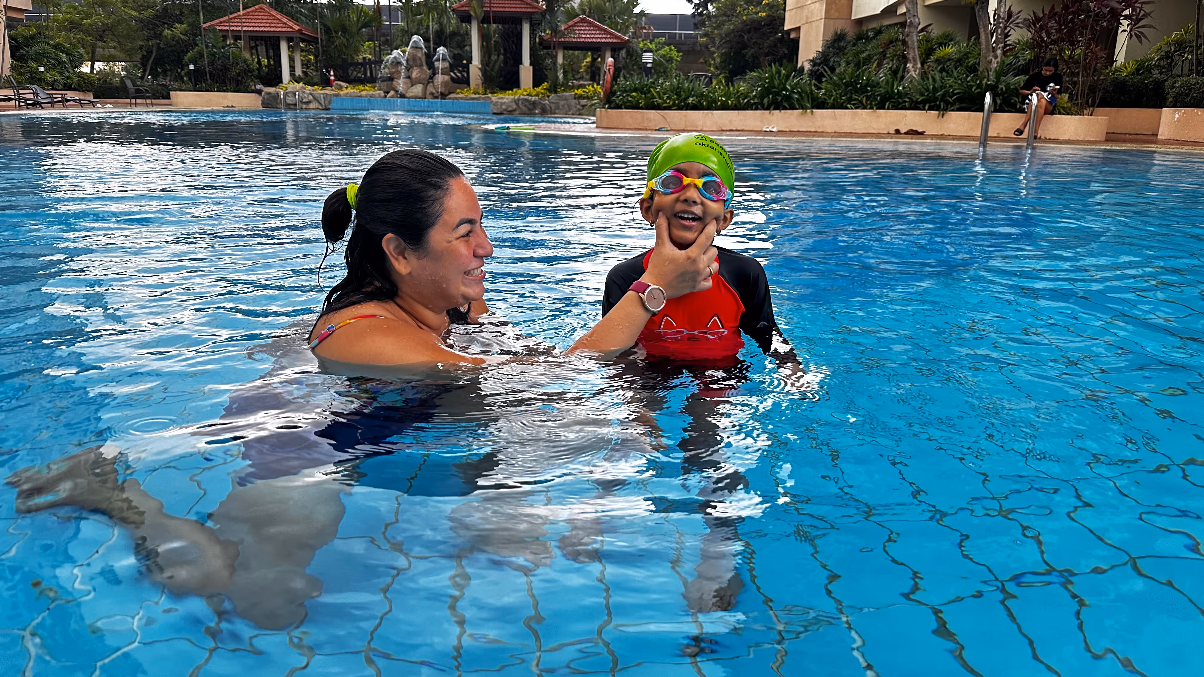 Woman smiling and holding a child wearing a green swim cap and goggles in a swimming pool.