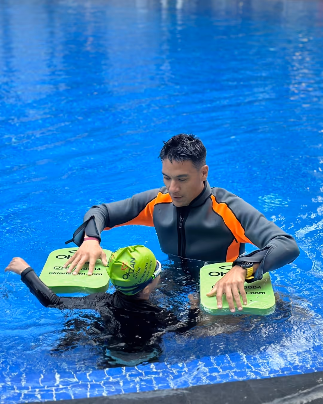 Man standing in a pool holding a smiling child wearing blue swimwear and goggles, both raising one arm in celebration.