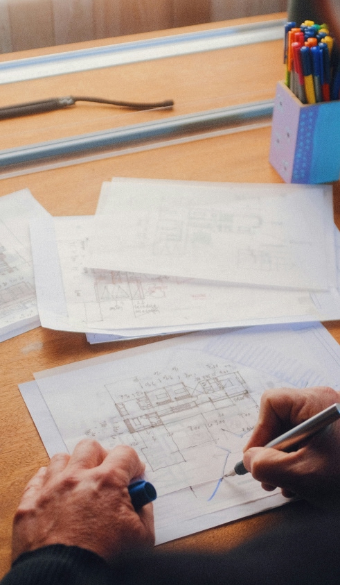Person drawing a blue line on architectural plans with a pen on a wooden desk with colored markers in a container.